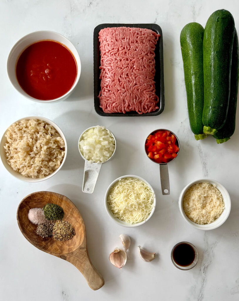 There are multiple sized bowls with ingredients in them: tomato sauce, cooked brown rice, shredded cheese, and Worcestershire sauce. There are 3 large green zucchinis, raw ground beef on a black tray, a measuring cup with chopped onion and another measuring cup with chopped red pepper. There is a large wooden spoon with dry spices in it and 3 garlic cloves. Items are on a white surface.