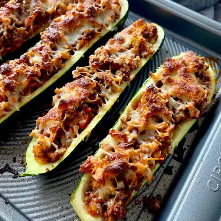 There is a large baking pan with baked hollowed out halved zucchinis filled with a ground beef and rice mixture. There is white shredded cheese sprinkled over the zucchini.