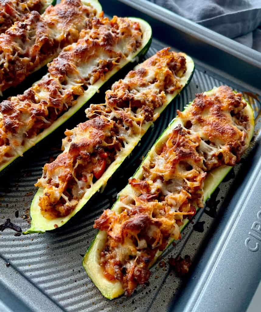 There is a large baking pan with baked hollowed out halved zucchinis filled with a ground beef and rice mixture. There is white shredded cheese sprinkled over the zucchini.