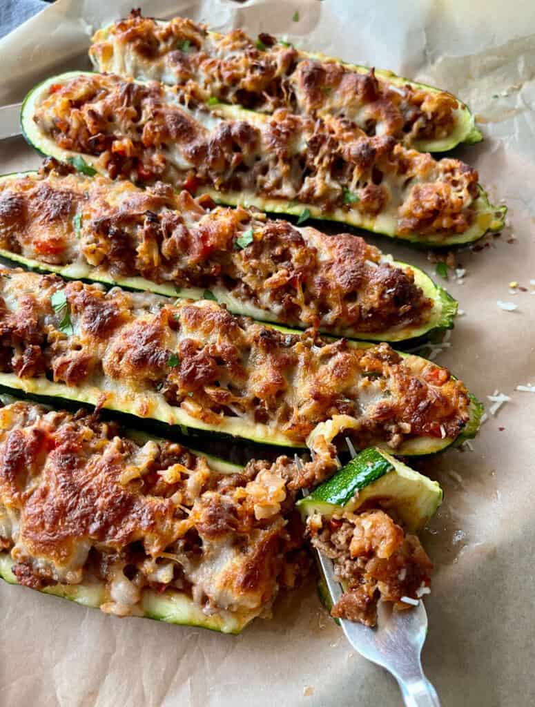 Baked hollowed out halved zucchinis filled with a ground beef and rice mixture. There is white shredded cheese sprinkled over the zucchini. The zucchini are on a wood tray lined with brown parchment paper. There is a fork with a piece of zucchini on it.