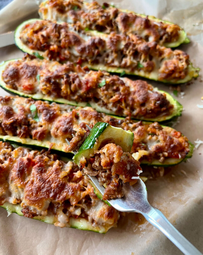 Baked hollowed out halved zucchinis filled with a ground beef and rice mixture. There is white shredded cheese sprinkled over the zucchini. The zucchini are on a wood tray lined with brown parchment paper. There is a fork with a piece of zucchini on it.