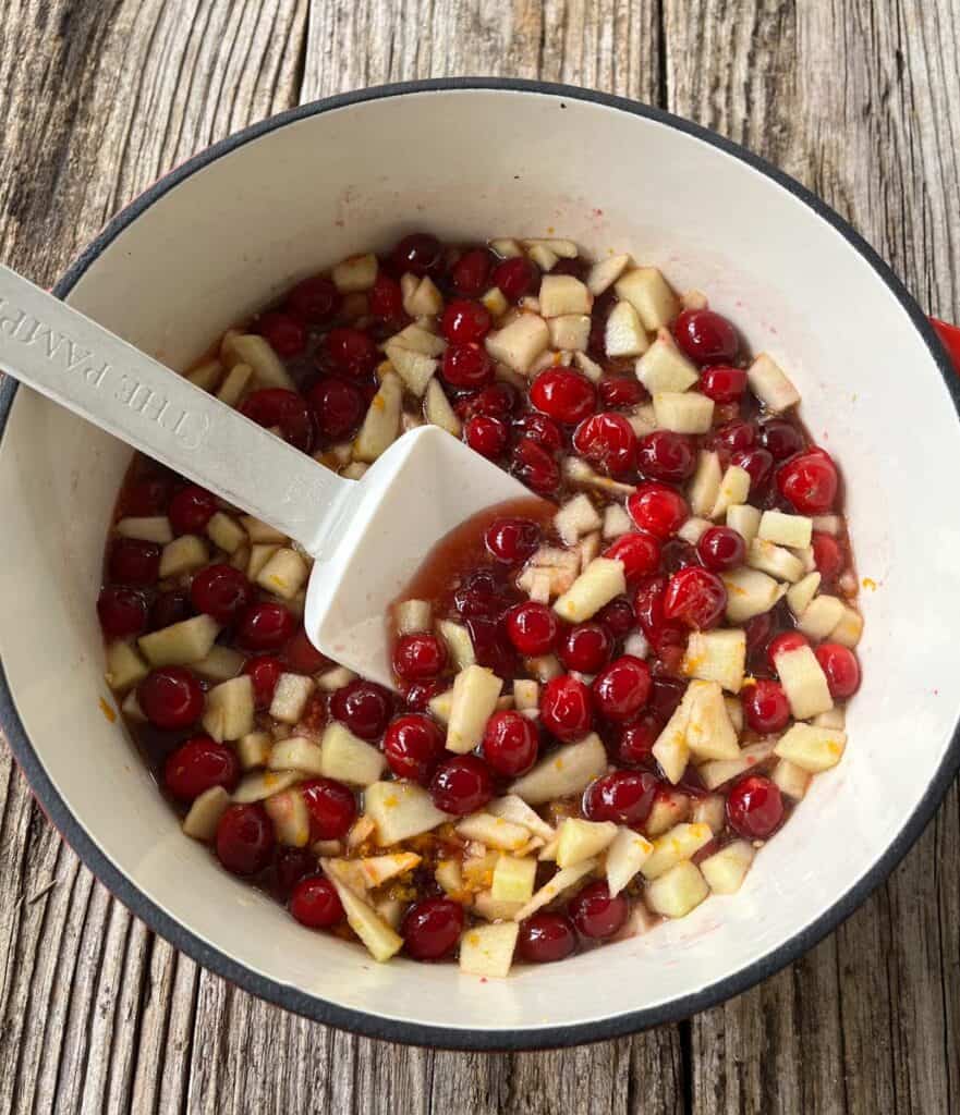 A pot of cranberries, orange peels, and apples with a spoon.