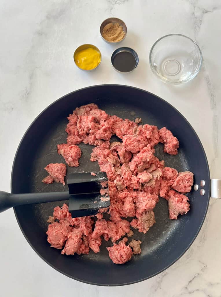 Large dark skillet with ground beef cooking. There is a black meat masher utensil resting in the skillet. There are small bowls. One with yellow mustard, one with brown sugar, and one with Worcestershire sauce. There is a small clear dish with water in it. Items are on a white surface.