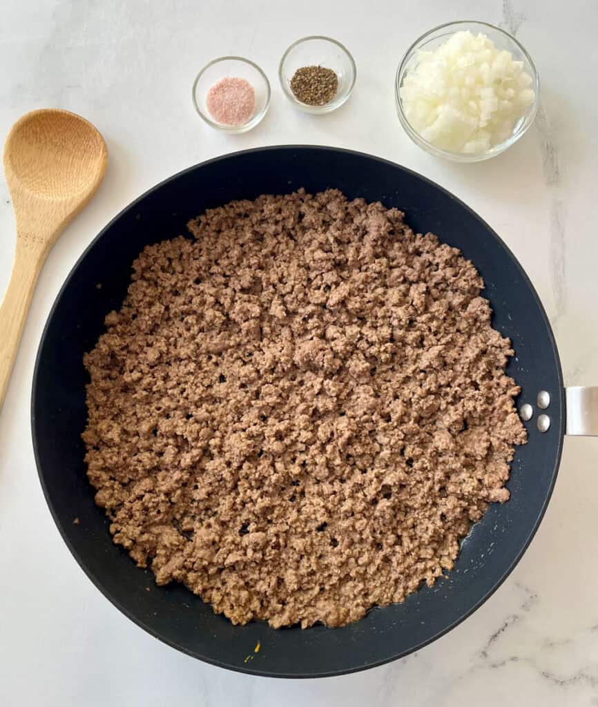 Large black skillet with a cooked ground beef mixture in it. There are small dishes with salt, pepper, and chopped onion in them. There is a wooden spoon . Items are on a white surface.