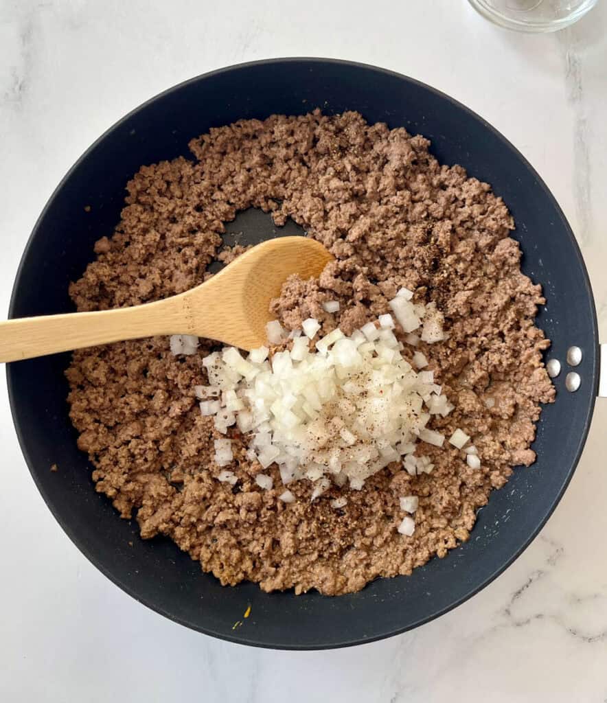 Large black skillet with a cooked ground beef mixture in it. There is a mound of chopped onion in the middle of the skillet. There is a wooden spoon resting in the skillet. The skillet is on a white surface.