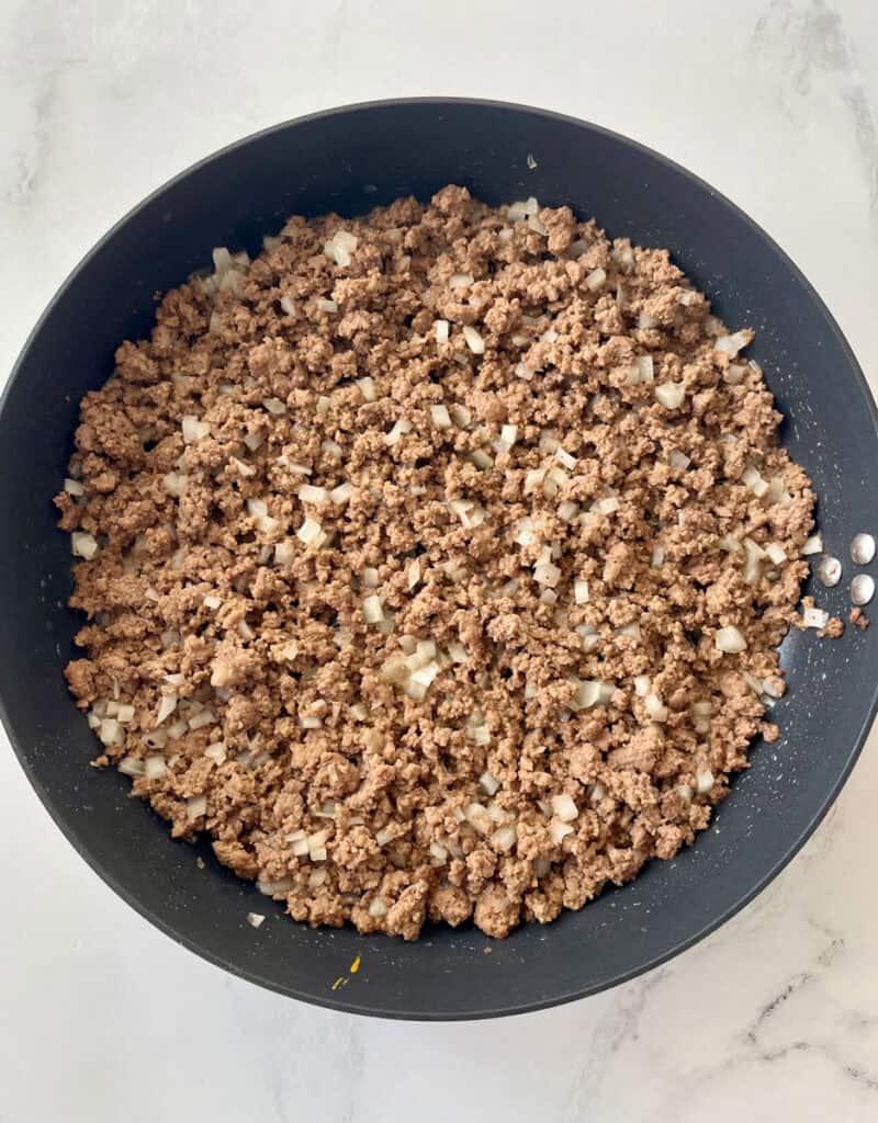 Large black skillet with a cooked ground beef mixture and chopped onion in it. Skillet is on a white surface.
