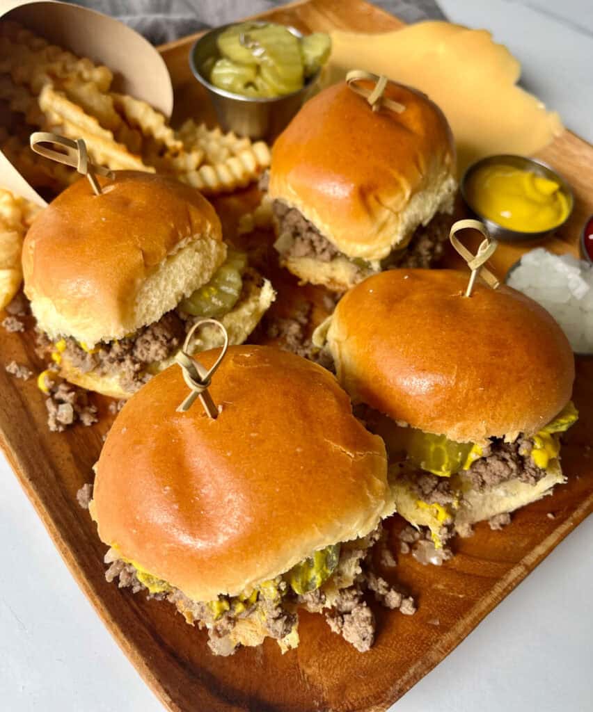 Large square platter with four small buns with a loose meat mixture, yellow mustard, and pickle slices in between the top and bottom. There are small wooden picks in each bun. There are crinkly french fries, a small dish with sliced pickles, a small dish with yellow mustard, a small dish with chopped onion, and slices of yellow cheese on the platter. Platter is on a white surface.