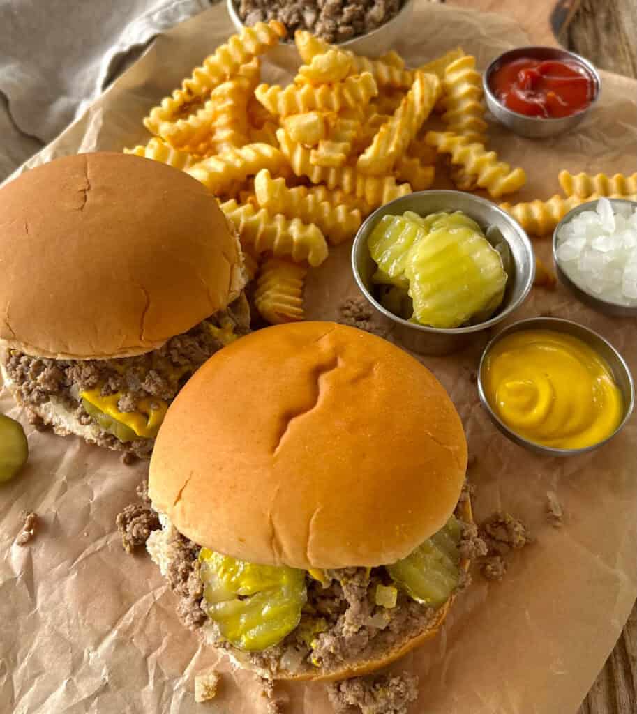 There are two hamburger buns with a loose meat mixture, pickle slices, and yellow mustard on them. There is a mound of crinkle french fries, a small dishes of mustard, of ketchup, of chopped onion, and one with sliced pickles. There is a small bowl of loose meat mixture. Items are on a wood surface that is lined with brown paper. There is a gray cloth napkin.