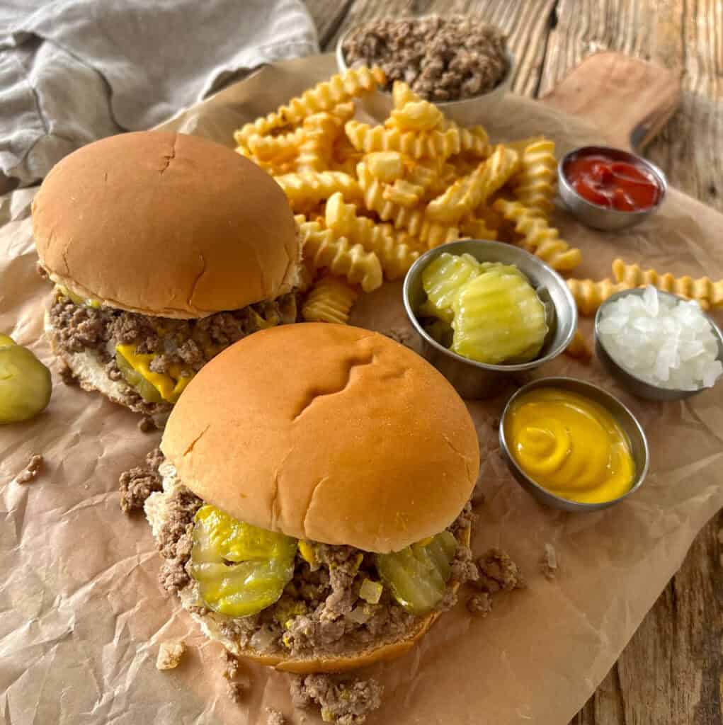 There are two hamburger buns with a loose meat mixture, pickle slices, and yellow mustard on them. There is a mound of crinkle french fries, a small dishes of mustard, of ketchup, of chopped onion, and one with sliced pickles. There is a small bowl of loose meat mixture. Items are on a wood surface that is lined with brown paper. There is a gray cloth napkin.