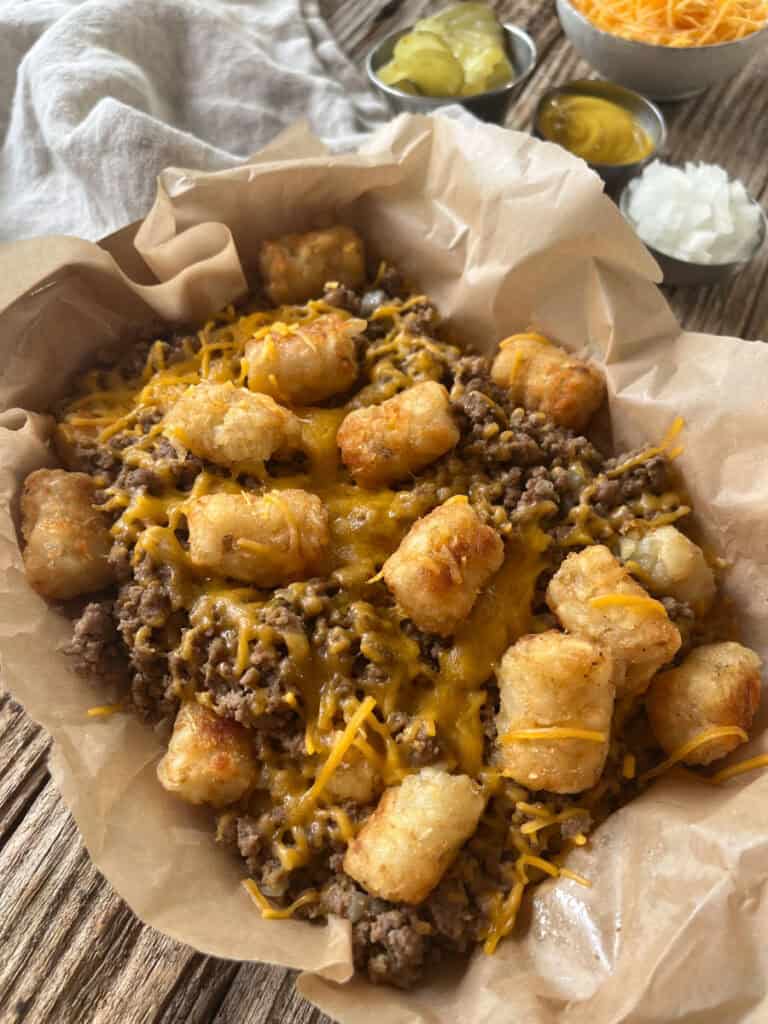 There is a loose ground meat mixture and small, cylindrical, deep fried potato pieces with melted cheese in a brown paper lined container. There are small dishes, one with yellow mustard, one with chopped onion, and one with shredded cheese. There is a light brown cloth napkin. Items are on a wood surface.