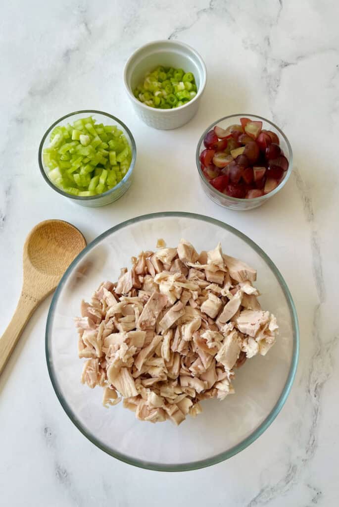 There is a large clear glass bowl with chopped cooked chicken in it. There are three small bowls, one with chopped celery, one with sliced green onions, and one with sliced red grapes. There is a wooden spoon laying next to the large glass bowl. Items are on a white surface.