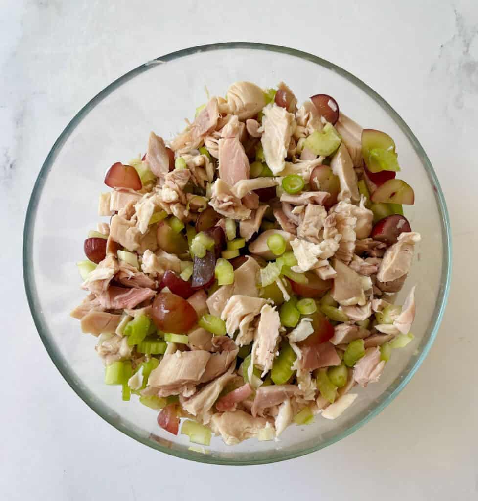 There is a large glass bowl with cooked chopped chicken, chopped celery, sliced green onions, and sliced red grapes mixed together. The bowl is on a white surface.