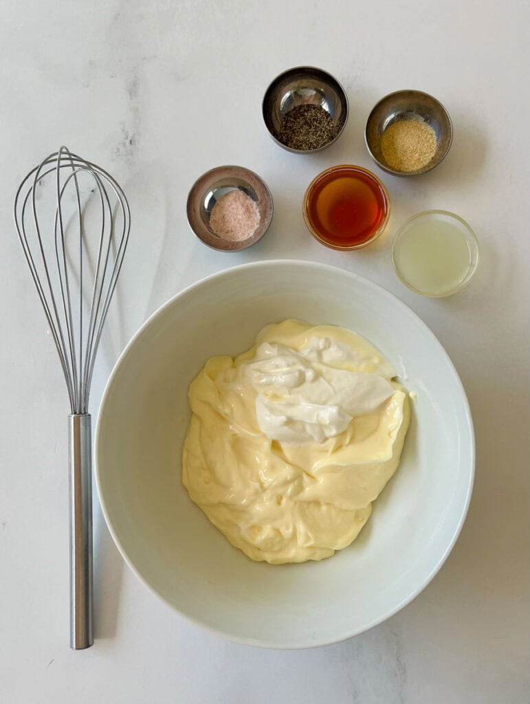 There is a large white bowl with mayonnaise and sour cream in it. There are five very small dishes, each with a separate ingredient: salt, pepper, onion powder, honey, and lemon juice. There is a metal whisk. Items are on a white surface.