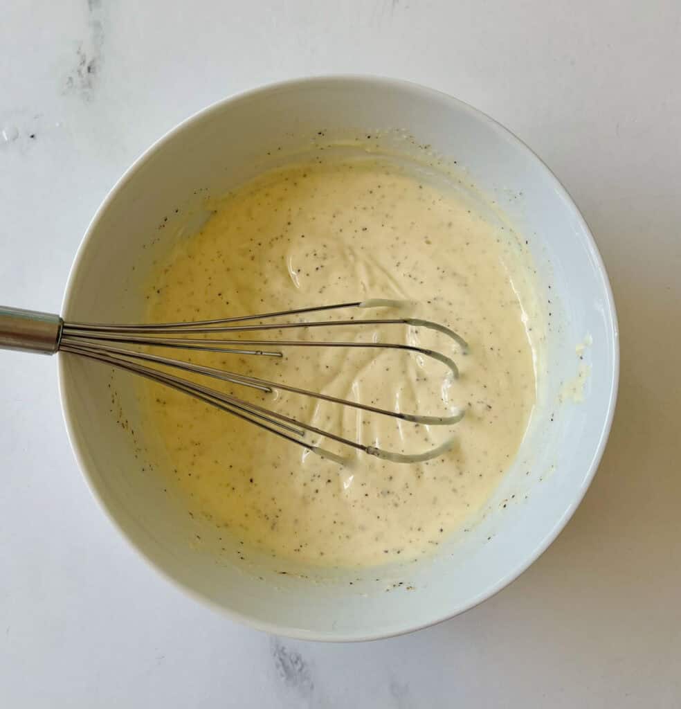 There is a large white bowl with a mixture of mayonnaise, sour cream, salt, pepper, onion powder, honey, and lemon juice. There is a metal whisk resting in the bowl. Bowl is on a white surface.