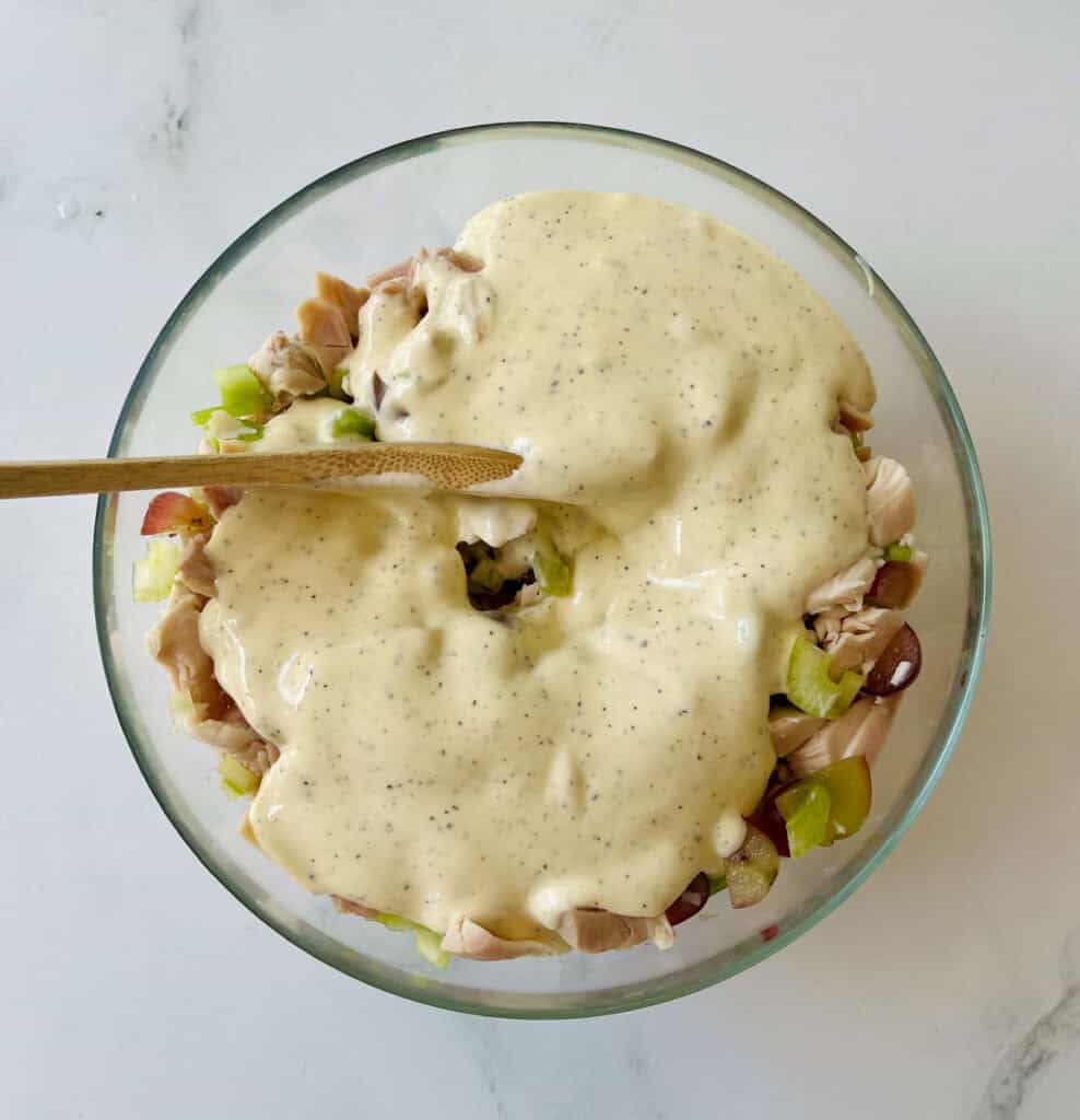 There is a large clear bowl with a mixture of chopped chicken, chopped celery, slice green onions, and sliced red grapes. There is a white sauce poured over the top. There is a wooden spoon in the bowl. The bowl is on a white surface.
