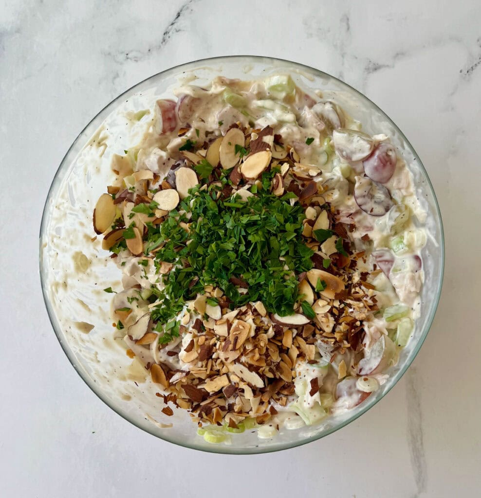 There is a large clear bowl with chicken salad in it. There is a mound of toasted sliced almonds and finely chopped parsley on top of the mixture. Bowl is on a white surface.