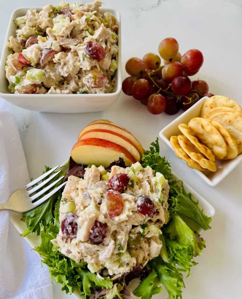 There is a white square plate with a scoop of chicken salad and apple slices on a bed of mixed greens. There is a fork resting on the plate. In the background there is a white bowl with chicken salad in it, red grapes, and a small white bowl with round crackers in it. There is a white cloth napkin next to the plate. Items are on a white surface.