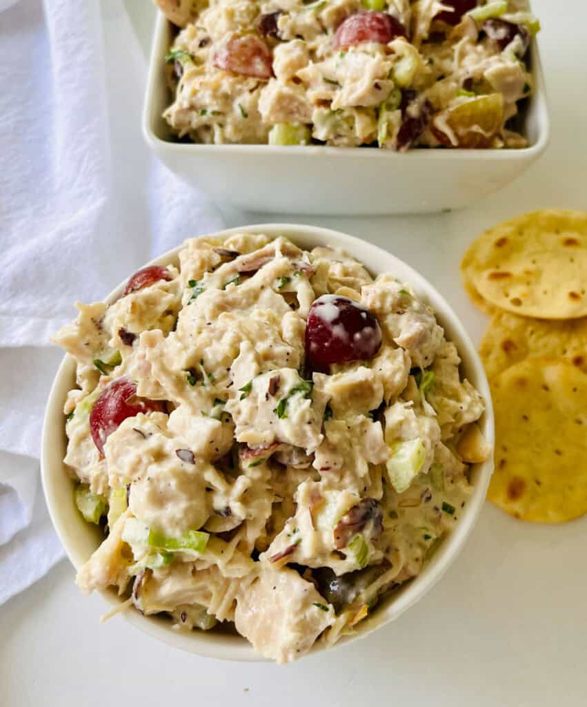 There is a white bowl with chicken salad. In the background there is a square bowl with chicken salad in it. There is a pile of round crackers and a white cloth napkin. Items are on a white surface.
