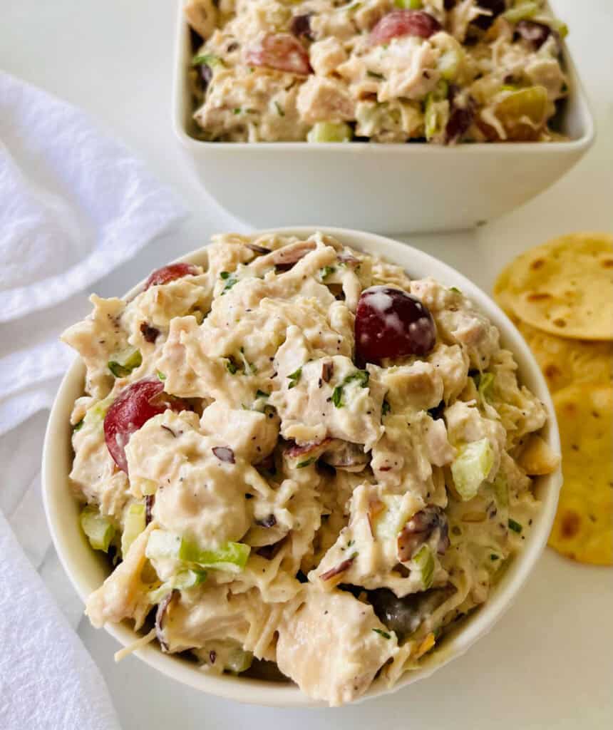 There is a white bowl with chicken salad. In the background there is a square bowl with chicken salad in it. There is a pile of round crackers and a white cloth napkin. Items are on a white surface.