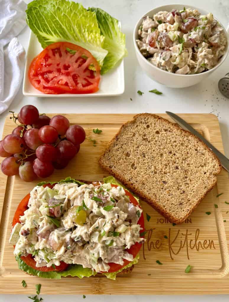 There is a rectangular wood board with two pieces of brown bread. On one of the pieces there is lettuce, sliced tomatoes and chicken salad. There is a small bunch of red grapes and a knife. In the background there is a white square plate with lettuce and sliced tomatoes on it. There is a white bowl with chicken salad in it. There is a white cloth napkin. Items are on a white surface.