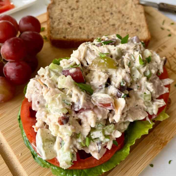 There is a rectangular wood board with two pieces of brown bread. On one of the pieces there is lettuce, sliced tomatoes and chicken salad. There is a small bunch of red grapes and a knife. Board is on a white surface.