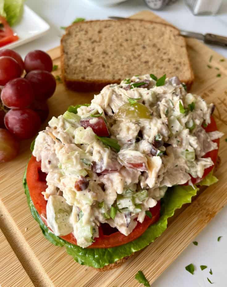 There is a rectangular wood board with two pieces of brown bread. On one of the pieces there is lettuce, sliced tomatoes and chicken salad. There is a small bunch of red grapes and a knife. Board is on a white surface.