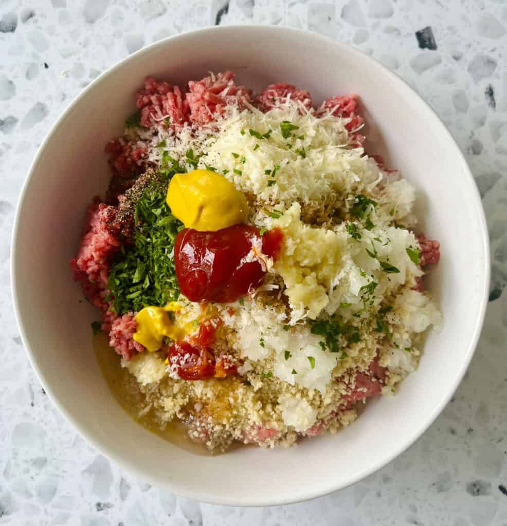 There is a large white bowl with raw ground beef, bread crumbs, egg, finely chopped onion, olive oil, grated parmesan cheese, fresh chopped parsley, Worcestershire sauce, yellow mustard, fresh minced garlic, ketchup, ground black pepper, and salt in it. Bowl is on a black and white terrazzo surface.