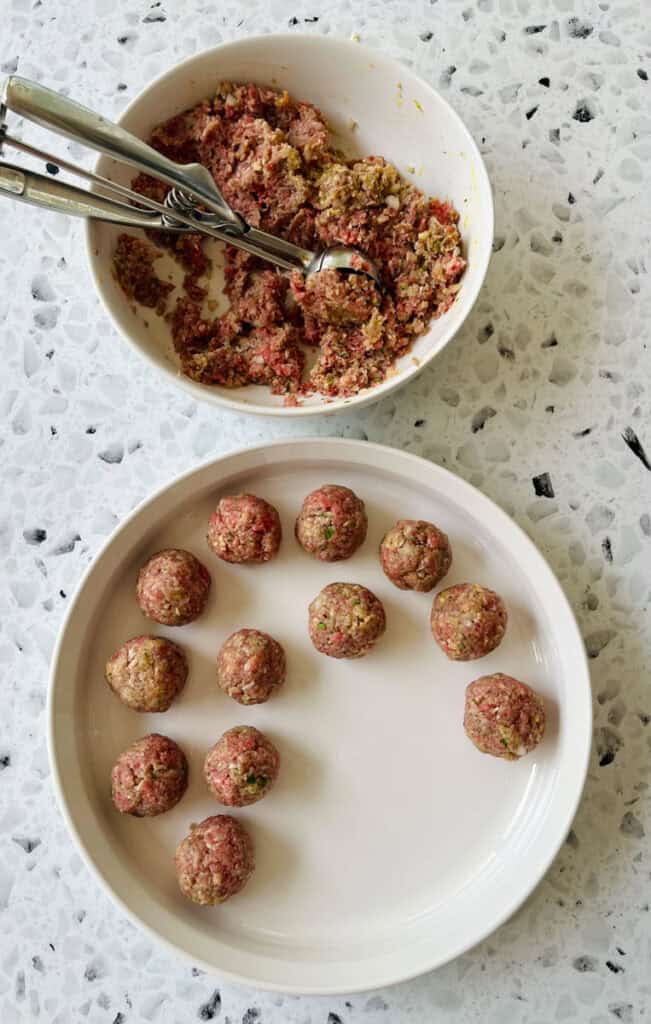 There is a large white bowl with a ground beef mixture in it. There is a scoop utensil resting in the bowl. There is a large white shallow round dish with meatballs in it. Bowls are on a black and white terrazzo surface.