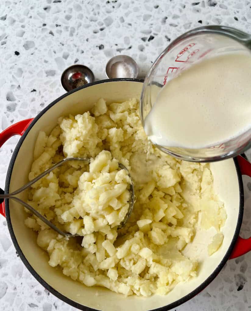 There is a large pot with cooked potato chunks. There is a masher utensil resting in the pot. There are two small dishes, one with salt and the other with pepper. There is a clear glass measuring cup with cream being poured into the pot. Items are on a black and white terrazzo surface.
