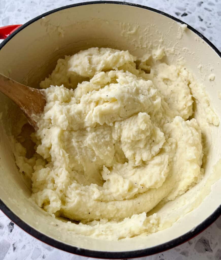 Large pot with mashed potatoes in it. There is a wooden spoon resting in the pot. Pot is on a black and white terrazzo surface.