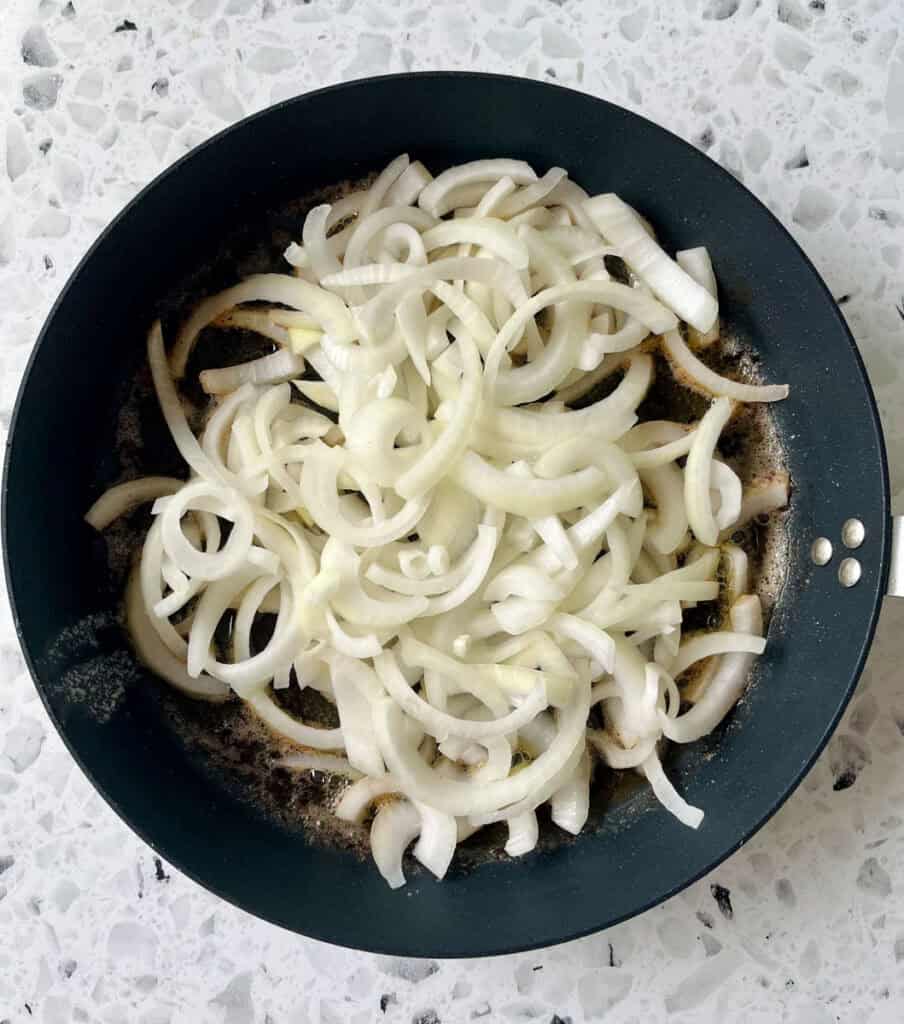 There is a large dark skillet with sliced onions cooking. Skillet is on a black and white terrazzo surface.
