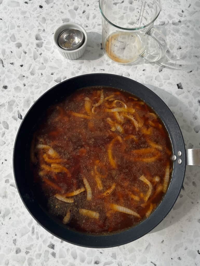 There is a large dark skillet with beef broth, cooked sliced onions and seasoning. There is an empty glass measuring cup and a stack of small bowls. Items are on a black and white terrazzo surface.