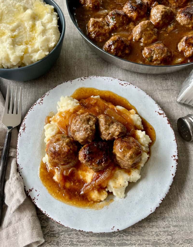 There is a plate with mashed potatoes, meatballs, and gravy on it. There is a tan cloth napkin and a fork next to the plate. There is a gray bowl with mashed potatoes, a dark skillet with meatballs and gravy, and a pair of salt and pepper shakers. Items are on a light gray surface.