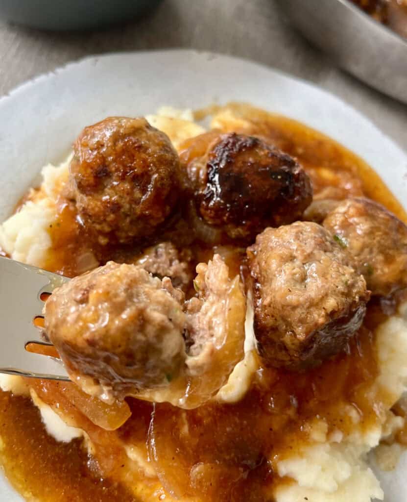 There is a plate with mashed potatoes, meatballs, and gravy on it. There is a fork with a meatball on it.