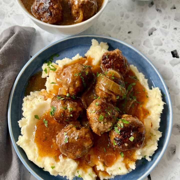 There is a blue plate with mashed potatoes, meatballs, and gravy on it. There is a gray cloth napkin laying next to the plate. In the background there are two white bowls. One bowl has mashed potatoes, the other has meatballs and gravy in it. Items are on a black and white terrazzo surface.