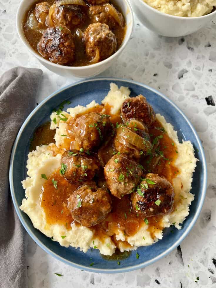 There is a blue plate with mashed potatoes, meatballs, and gravy on it. There is a gray cloth napkin laying next to the plate. In the background there are two white bowls. One bowl has mashed potatoes, the other has meatballs and gravy in it. Items are on a black and white terrazzo surface.