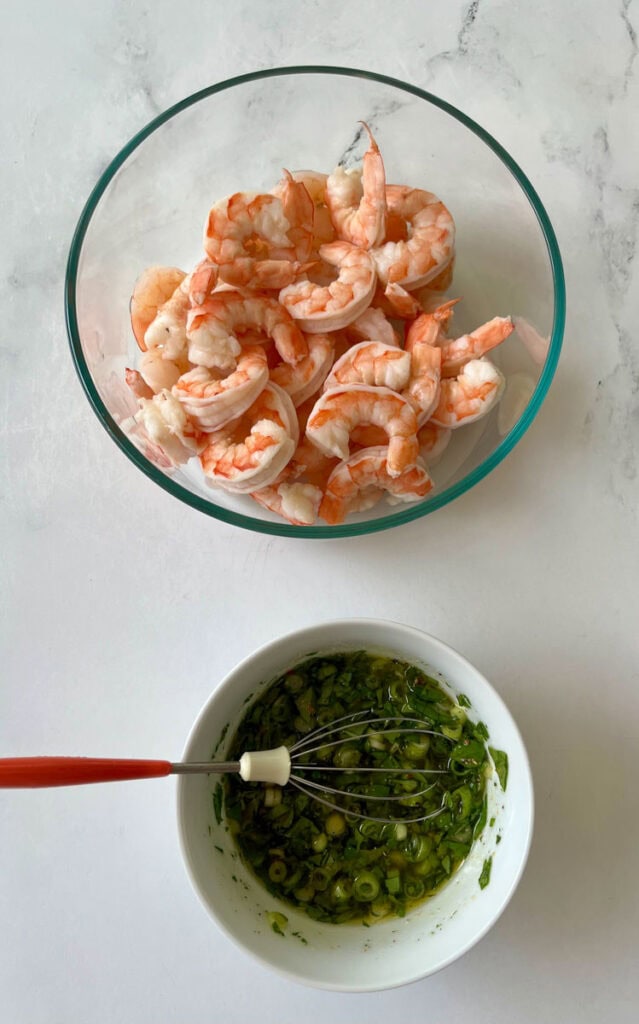 There is a large clear glass bowl with cooked shrimp and a small white bowl with a marinade of olive oil, white vinegar, sliced green onion, chopped fresh herbs and dry seasonings. There is a small metal whisk resting in the bowl. Items are on a white surface.