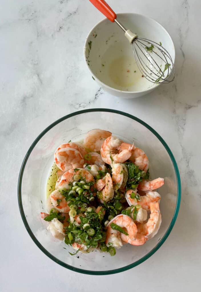 There is a large clear glass bowl with cooked shrimp and a marinade of olive oil, white vinegar, sliced green onion, chopped fresh herbs and dry seasonings. There is an empty small bowl with a small metal whisk resting in the bowl. Items are on a white surface.