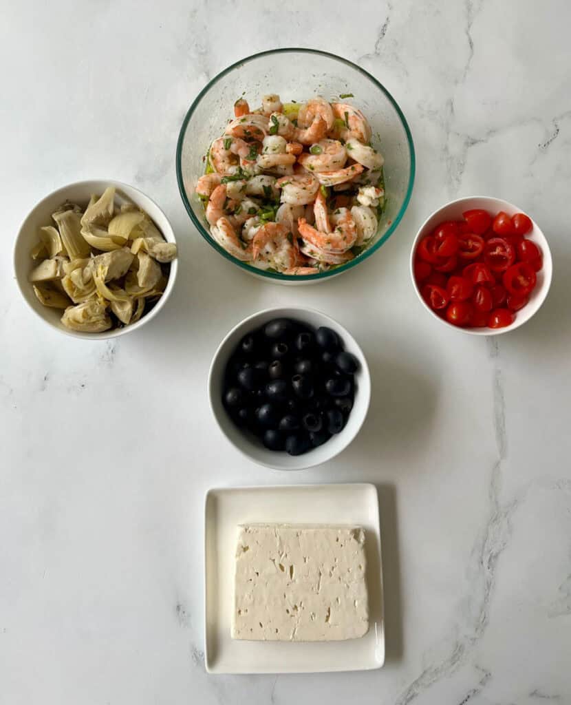 There is a large clear glass bowl with cooked shrimp and a marinade of olive oil, white vinegar, sliced green onion, chopped fresh herbs and dry seasonings mixed together. There are three small bowls, one with chopped artichoke hearts, one with whole black olives, and one with halved small tomatoes. There is a square white plate with a block of white feta cheese on it. Items are on a white surface.