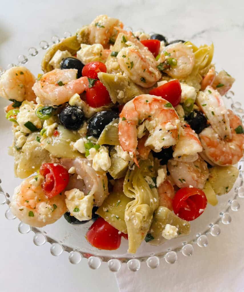 There is a large clear bowl with marinated shrimp, black olives, crumbled feta cheese, chopped artichoke hearts, and small tomatoes cut in half. Bowl is on a white surface.