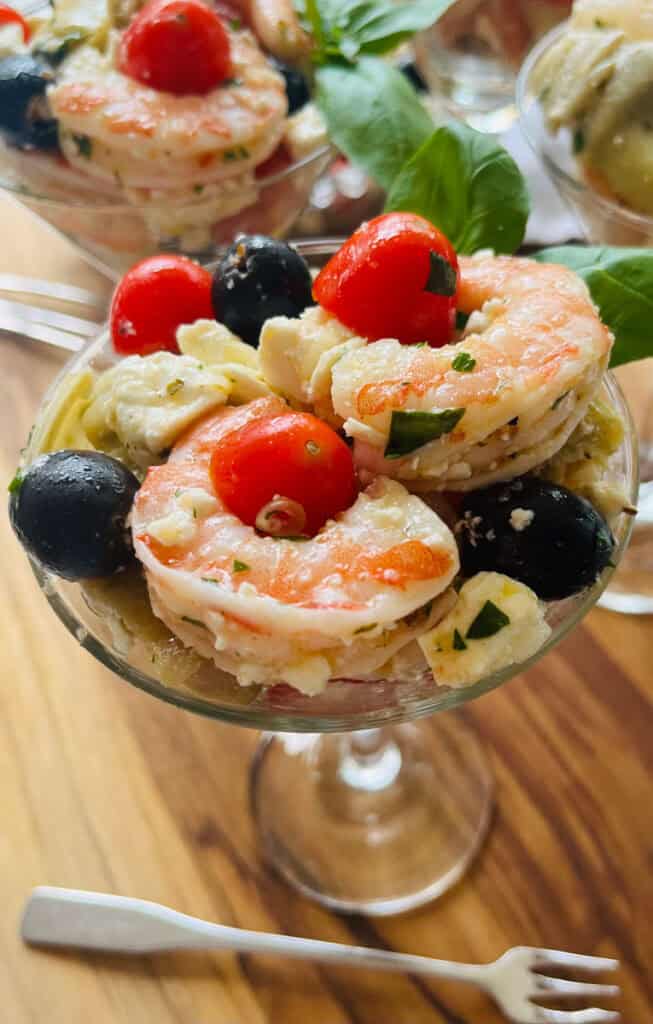 There are small clear stemmed glasses with with marinated shrimp, black olives, crumbled feta cheese, chopped artichoke hearts, and small tomatoes cut in half in them. There is a small fork laying next to one of the glasses. Glasses are on a wood surface.