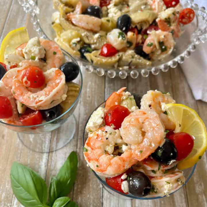 There are two small clear stemmed glasses and a large clear bowl with marinated shrimp, black olives, crumbled feta cheese, chopped artichoke hearts, and small tomatoes cut in half in them. There is a white cloth napkin. Items are on a wood surface.