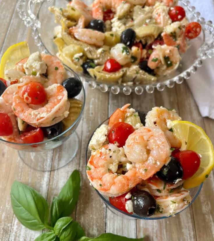 There are two small clear stemmed glasses and a large clear bowl with marinated shrimp, black olives, crumbled feta cheese, chopped artichoke hearts, and small tomatoes cut in half in them. There is a white cloth napkin. Items are on a wood surface.