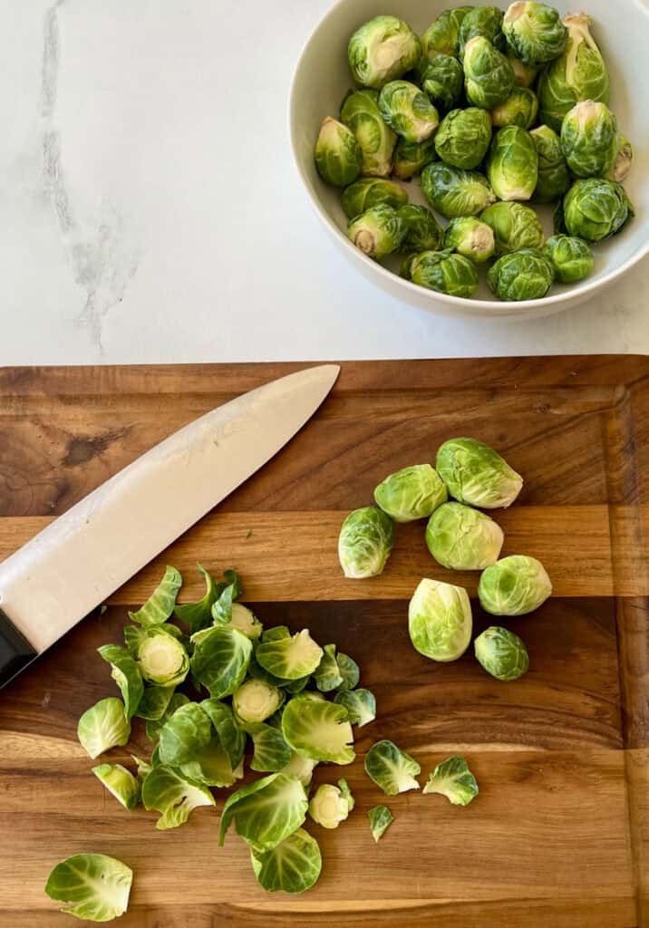 There is a large wooden board with Brussels sprouts and a large cutting knife on it. There is a large white bowl with Brussels sprouts in it in the background. Items are on a white surface.