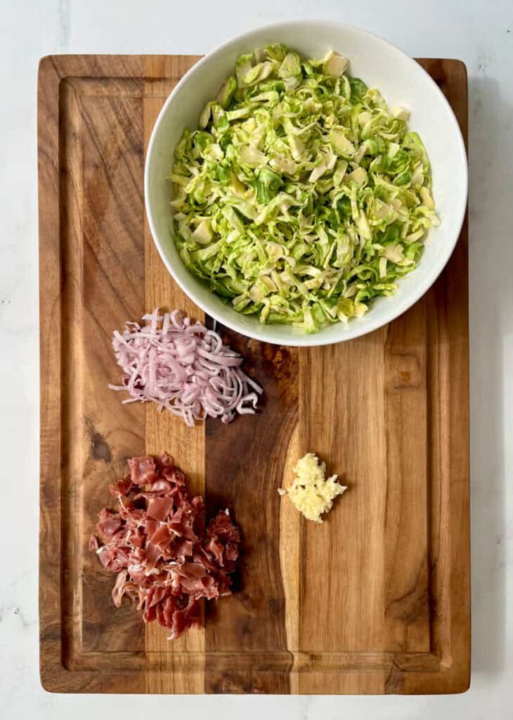 There is a large wooden board with a bowl of shredded Brussels sprouts, a mound of sliced onions, a mound of chopped prosciutto, and minced garlic on it. Board is on a white surface.