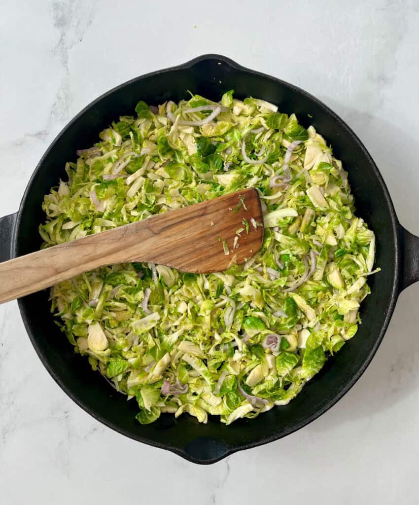 Large black skillet with shredded Brussels sprouts and sliced onions in it. There is a wooden spoon resting in the skillet. Skillet is on a white surface.