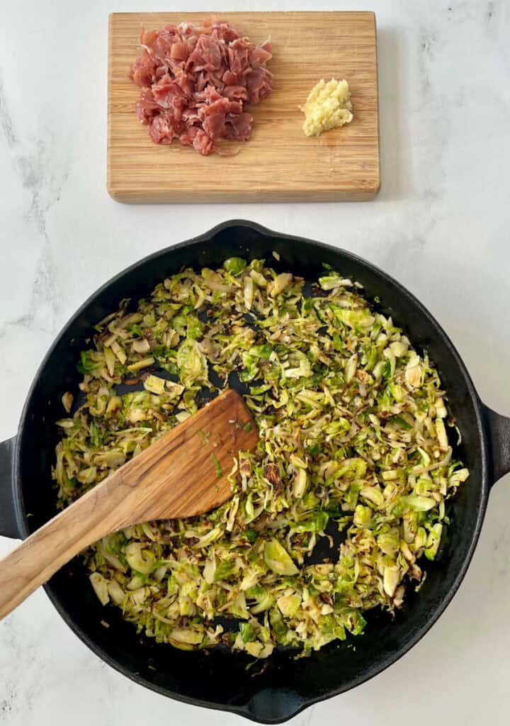 Large black skillet with cooked shredded Brussels sprouts and sliced onions. There is a wooden spoon resting in the skillet. There is a wood board with chopped prosciutto and minced garlic on it in the background. Items are on a white surface.