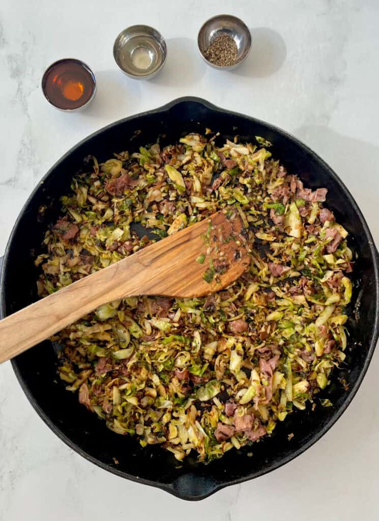 Large black skillet with cooked shredded Brussels sprouts, pieces of prosciutto, and seasonings. There is a wooden spoon resting in the skillet. There are three small ramekins with seasonings in the background. Items are on a white surface.