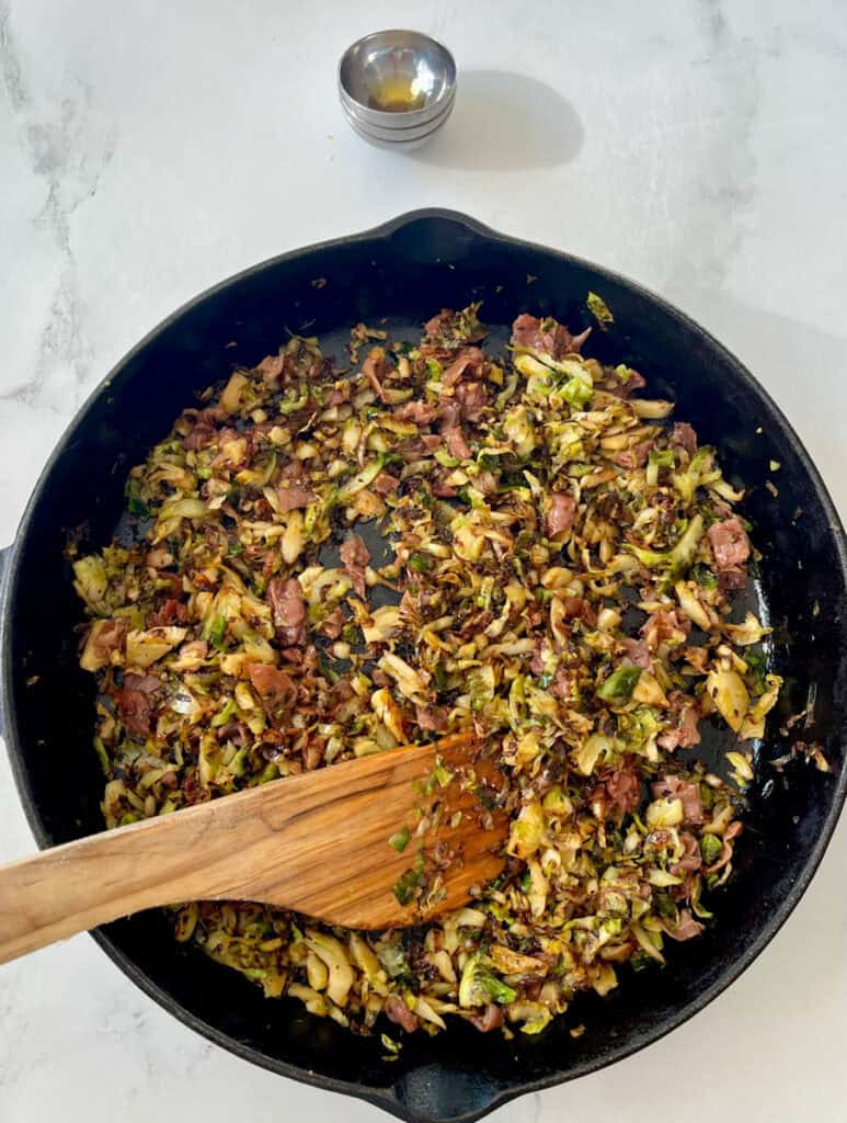 Large black skillet with cooked shredded Brussels sprouts, pieces of prosciutto, and seasonings. There is a wooden spoon resting in the skillet. There is a stack of empty ramekins in the background. Items are on a white surface.
