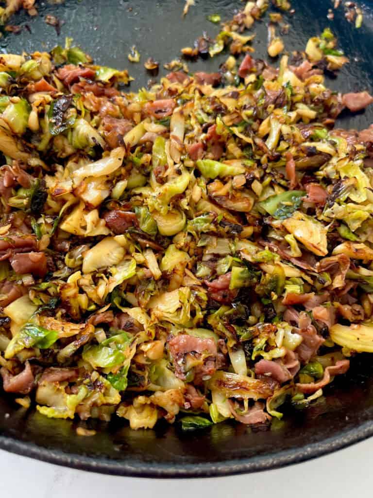Large black skillet with cooked shredded Brussels sprouts, pieces of prosciutto, and seasonings.